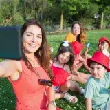 A joyful family picnic with a woman and children taking a group selfie in a sunny park.