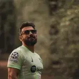 A bearded man in a soccer jersey stands confidently in a serene forest setting.