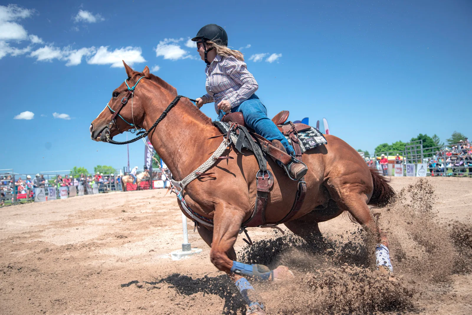 A woman rides a horse performing a fast turn at a rodeo under a bright blue sky.
