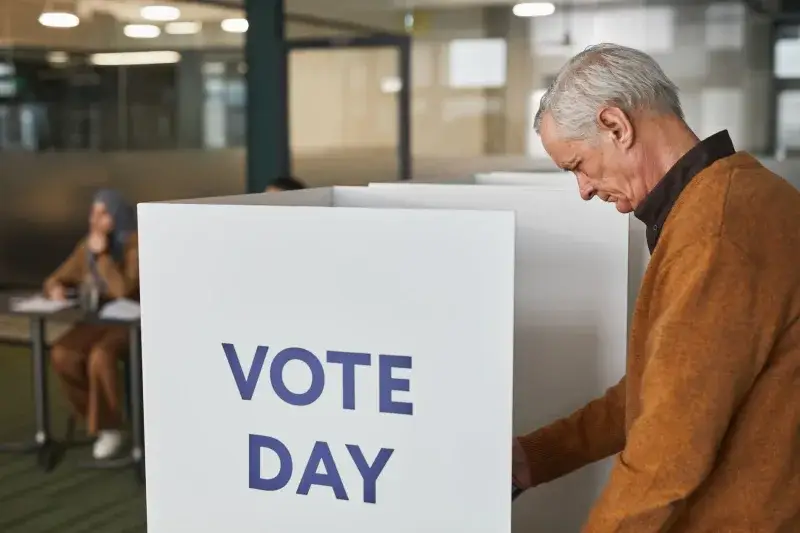 Senior man in brown sweater voting at an indoor polling station on election day.