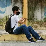 A man wearing a mask sits on the street pavement against a post in urban Venezuela.