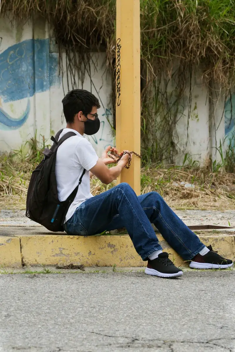 A man wearing a mask sits on the street pavement against a post in urban Venezuela.