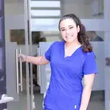 Smiling female nurse in blue scrubs opens a glass door in a healthcare setting.