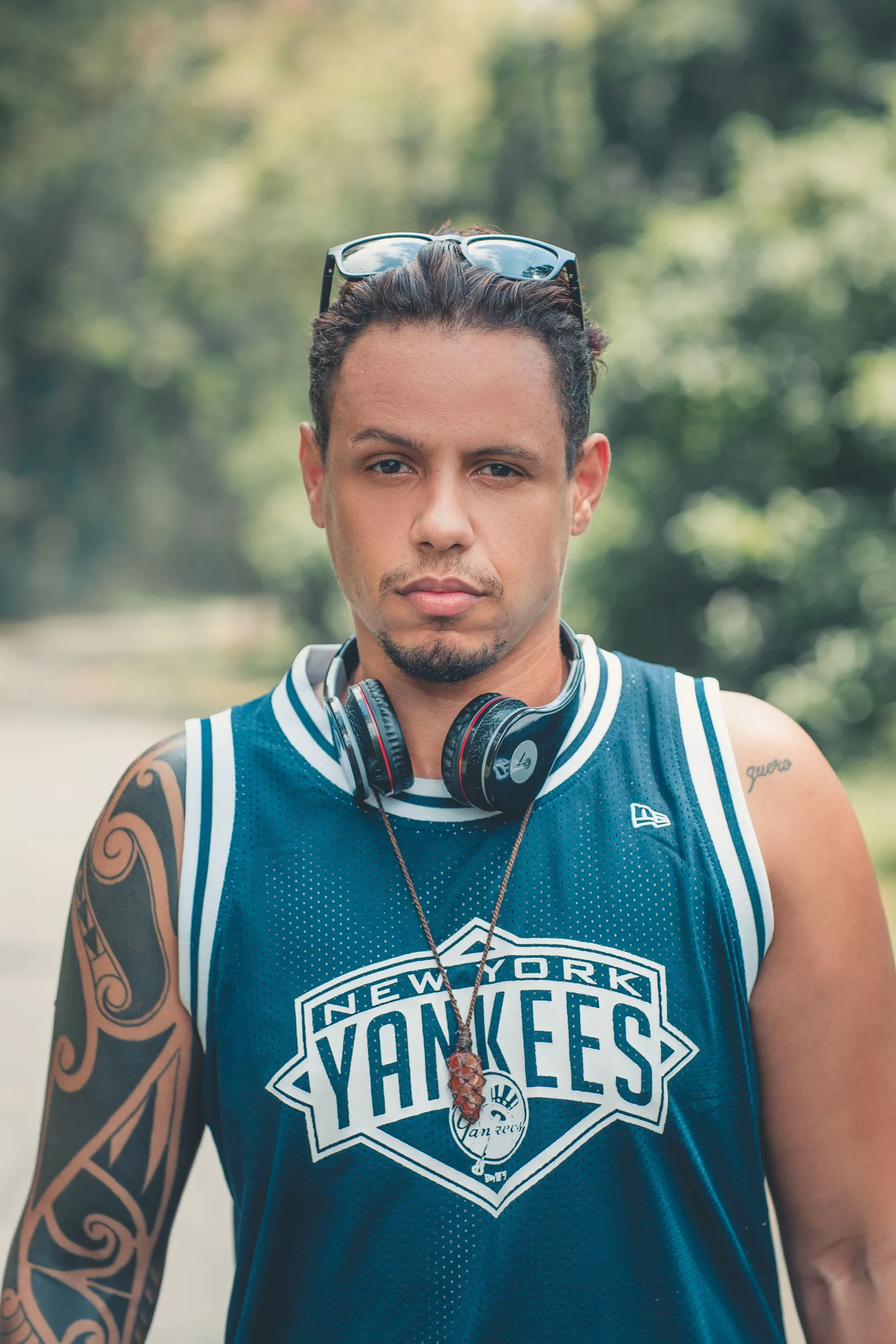 Un hombre de aspecto serio con tatuajes y auriculares en un entorno al aire libre, vistiendo una camiseta de los New York Yankees.