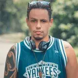 Un hombre de aspecto serio con tatuajes y auriculares en un entorno al aire libre, vistiendo una camiseta de los New York Yankees.