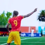 Football player in red and yellow kit gestures on field during outdoor match.