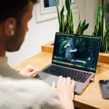 A man using a laptop for photo editing in a modern office environment, with plants and a smartphone nearby.