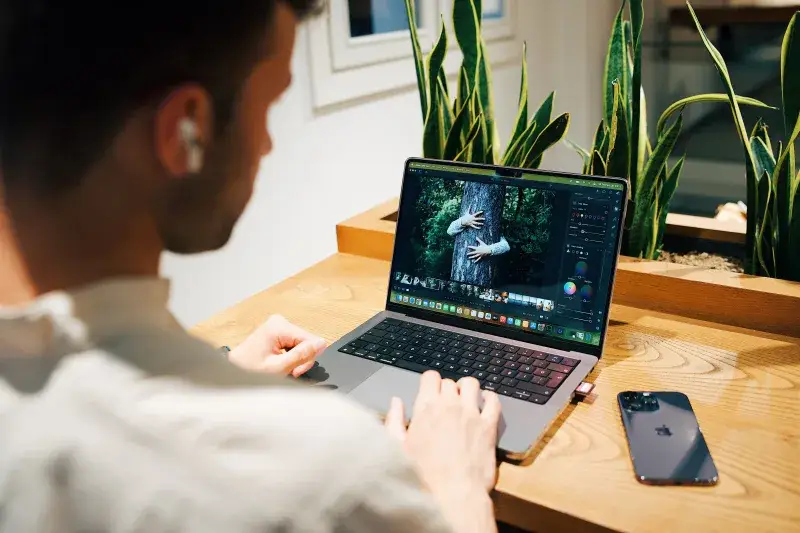 A man using a laptop for photo editing in a modern office environment, with plants and a smartphone nearby.