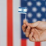 Closeup of crop unrecognizable person holding small flag of Israel before huge flag of United States of America on background