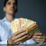 A man in a white shirt holds a fan of Argentine peso banknotes, representing wealth and finance.