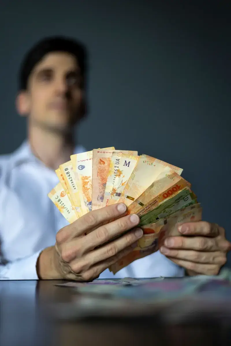 A man in a white shirt holds a fan of Argentine peso banknotes, representing wealth and finance.