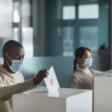 People wearing face masks cast ballots in a public indoor polling station with social distancing.
