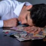 A man in a white shirt rests his head on a table covered with Argentine pesos and coins.