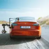 Red Tesla Model 3 with a camera rig, driving on a sandy road under a clear sky.