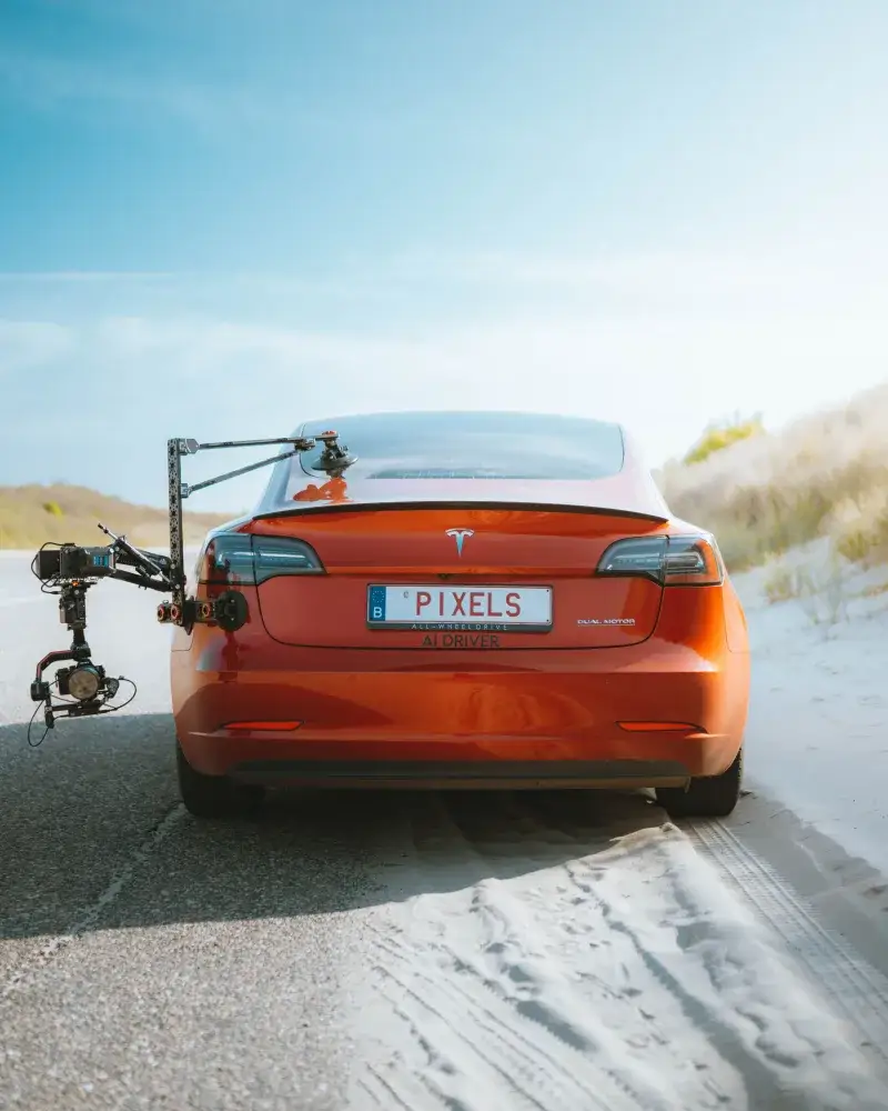 Red Tesla Model 3 with a camera rig, driving on a sandy road under a clear sky.