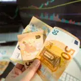Hand holding Euro banknotes in front of a trading screen showing financial graphs.