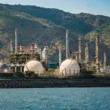 Industrial refinery with spherical tanks by the coastline against a backdrop of mountains.