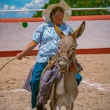 A woman in traditional attire rides a donkey at a cultural rodeo in Pachuca, Mexico.