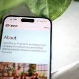 Close-up shot of a smartphone screen showing the OpenAI website with greenery in the background.