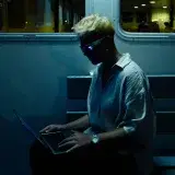 A person with platinum blonde hair uses a laptop on a ferry in İstanbul at night.