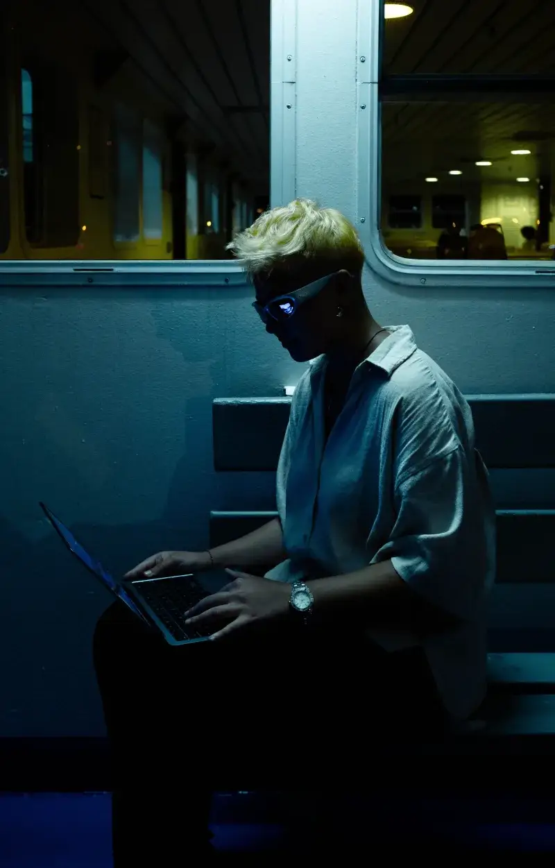 A person with platinum blonde hair uses a laptop on a ferry in İstanbul at night.