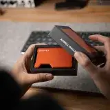 Hands holding an orange metal wallet during unboxing in an indoor office environment.
