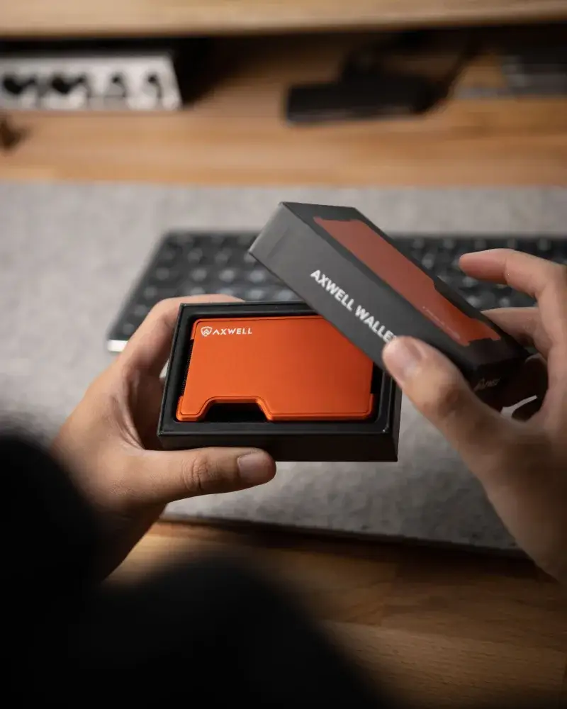 Hands holding an orange metal wallet during unboxing in an indoor office environment.