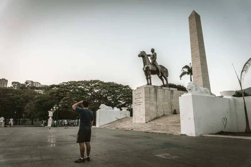 A man stands near a historic statue and obelisk in a plaza in Caracas, Venezuela.