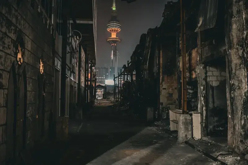 Night view of the Kuwait Tower brightly illuminated, standing amidst dark, decaying urban surroundings.
