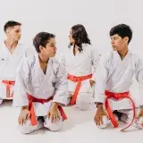 Group of teenagers in karate uniforms with red belts sitting in a studio setting.