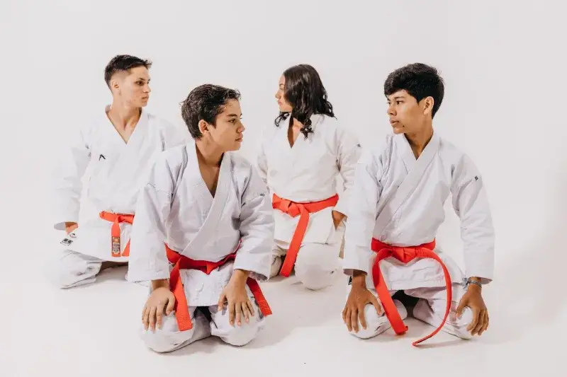 Group of teenagers in karate uniforms with red belts sitting in a studio setting.