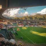A bustling soccer stadium at sunset with spectators enjoying the game amidst scenic mountains.