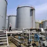 Persona caminando cerca de grandes tanques de almacenamiento de petróleo industriales y tuberías bajo un cielo azul claro.