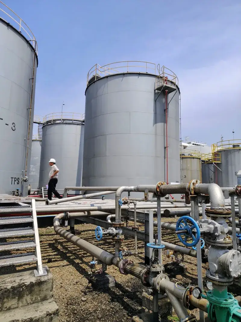 Persona caminando cerca de grandes tanques de almacenamiento de petróleo industriales y tuberías bajo un cielo azul claro.