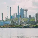 A large industrial refinery complex with tall chimneys by the waterfront under a clear sky.