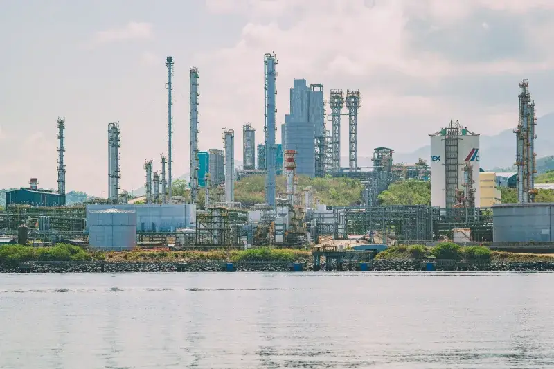 A large industrial refinery complex with tall chimneys by the waterfront under a clear sky.