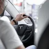 Interior view of a person driving a Tesla vehicle, with focus on the steering wheel and dashboard.