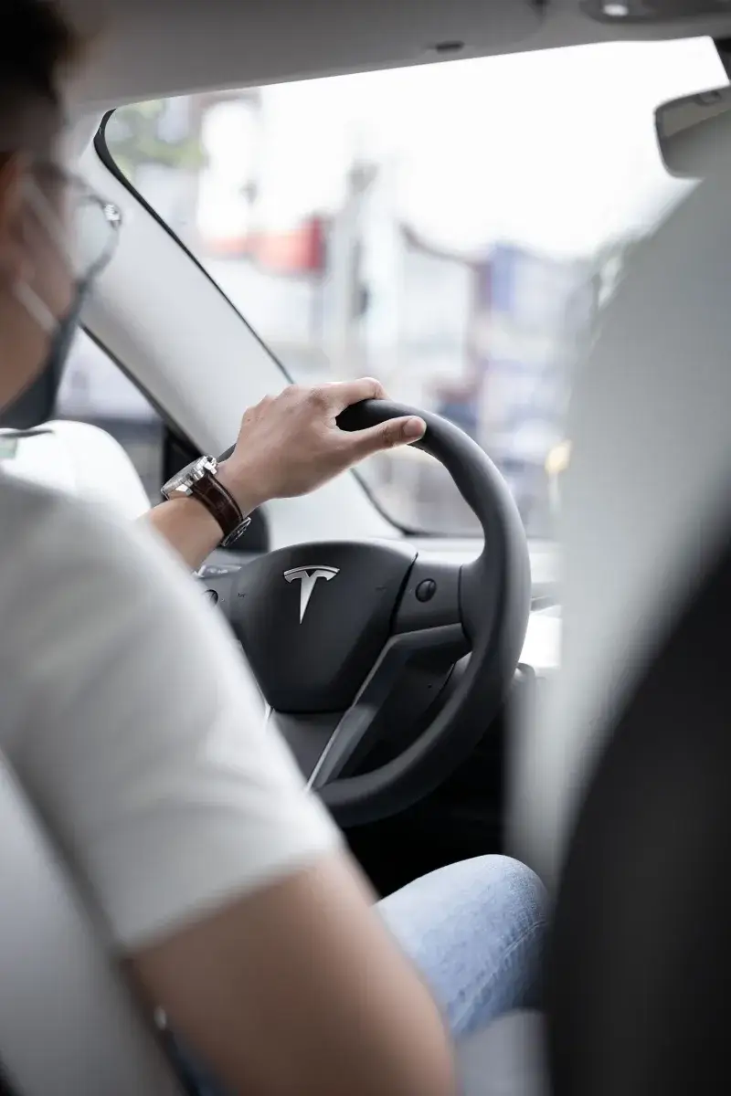 Interior view of a person driving a Tesla vehicle, with focus on the steering wheel and dashboard.