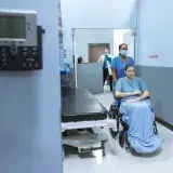 A nurse helps a patient in a wheelchair down a hospital corridor, reflecting care and medical professionalism.