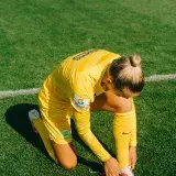 A female soccer player in yellow sportswear tying her shoelaces on a grass field.