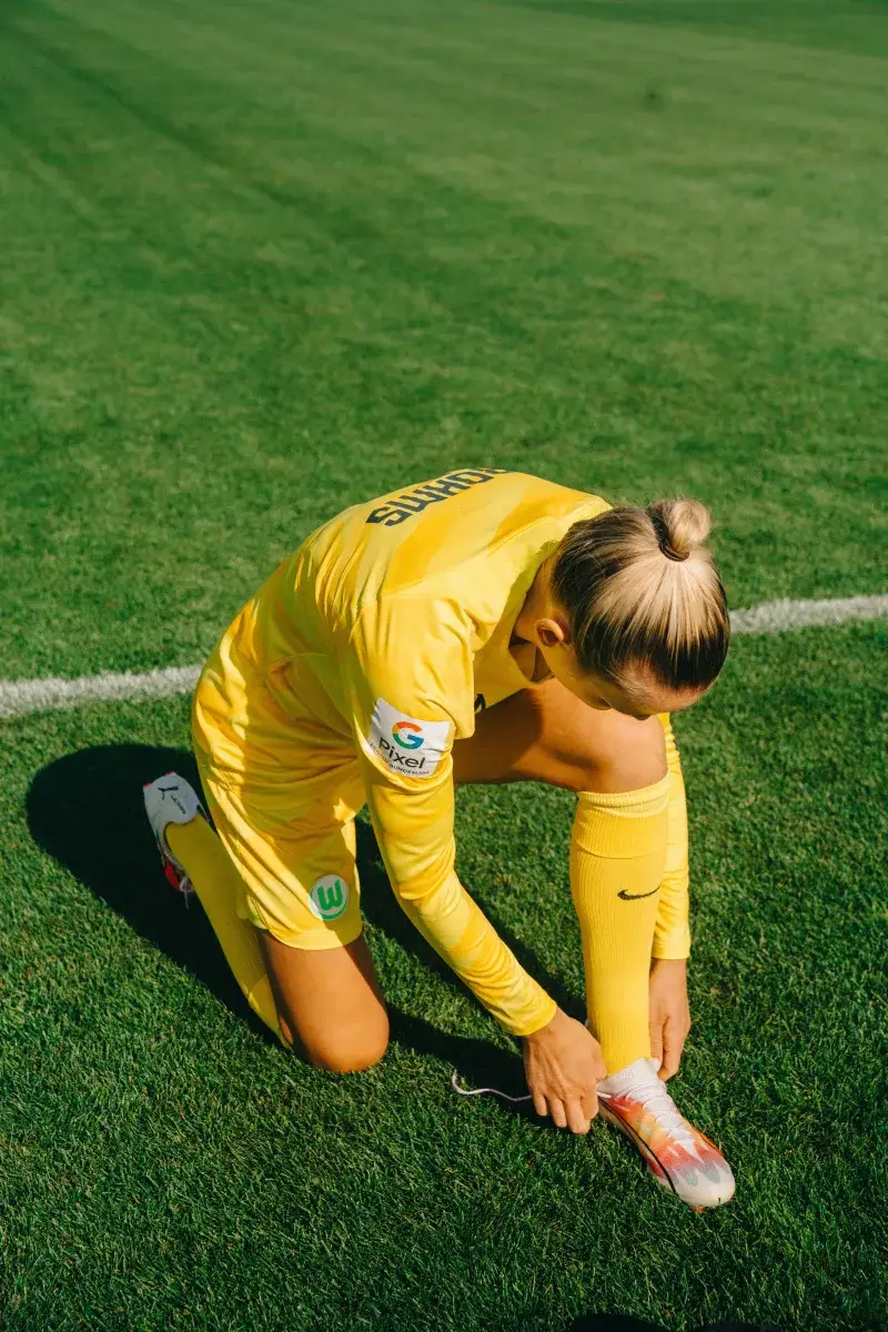 A female soccer player in yellow sportswear tying her shoelaces on a grass field.