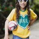 Cheerful child with curly hair wearing a Brasil shirt and sunglasses, holding a soccer ball outdoors.