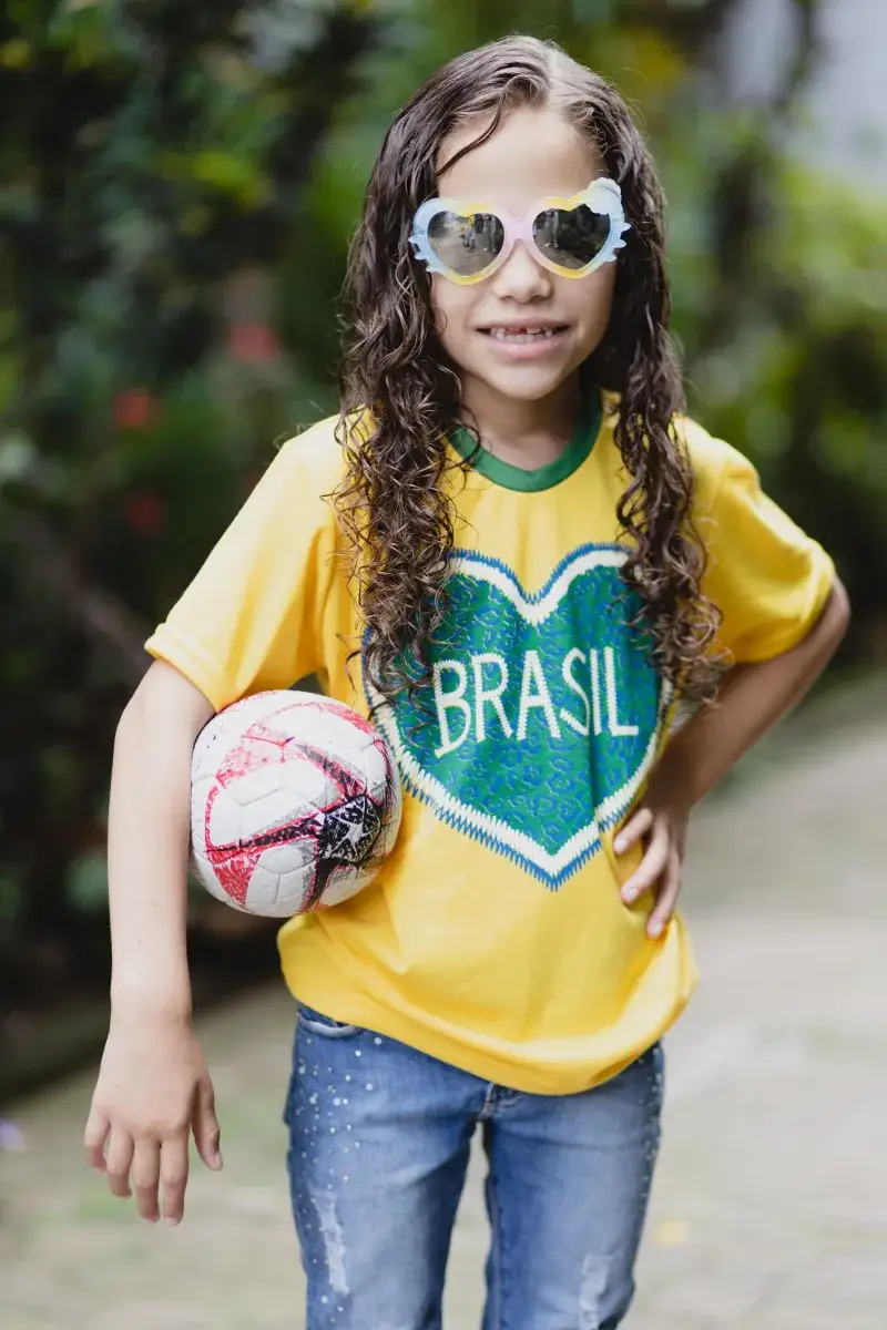 Cheerful child with curly hair wearing a Brasil shirt and sunglasses, holding a soccer ball outdoors.