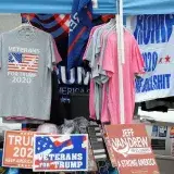 A stall displaying Trump 2020 merchandise including shirts and signs at an outdoor market.