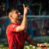 A football player in a red jersey celebrating with a finger point during a match outdoors.