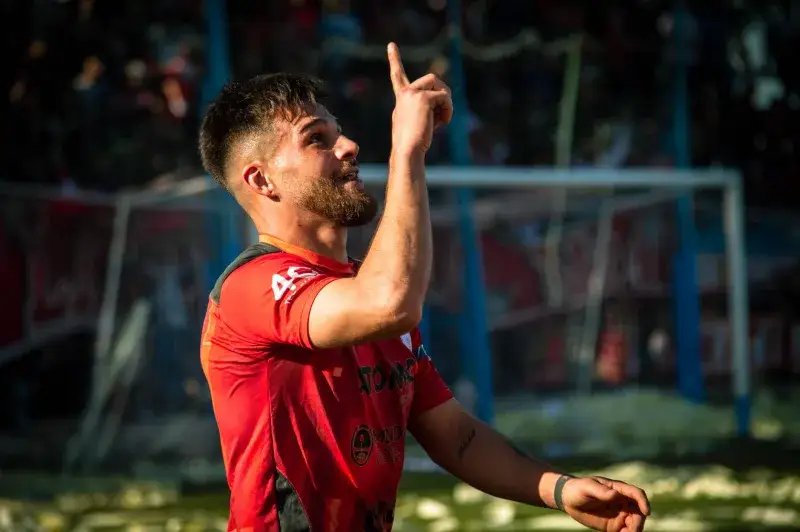 A football player in a red jersey celebrating with a finger point during a match outdoors.
