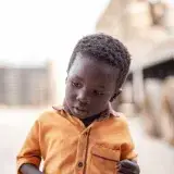 A young boy in an orange shirt wandering outdoors in Sampa, Bono Region, Ghana.