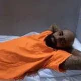 Man in orange prison uniform lying on bed, showing tattoos, indoors setting.