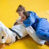 Two adults practicing judo in a grappling hold on a yellow mat indoors.