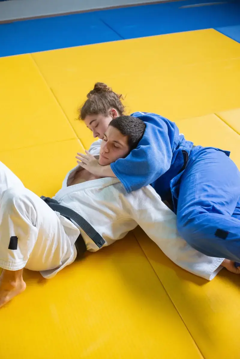 Two adults practicing judo in a grappling hold on a yellow mat indoors.